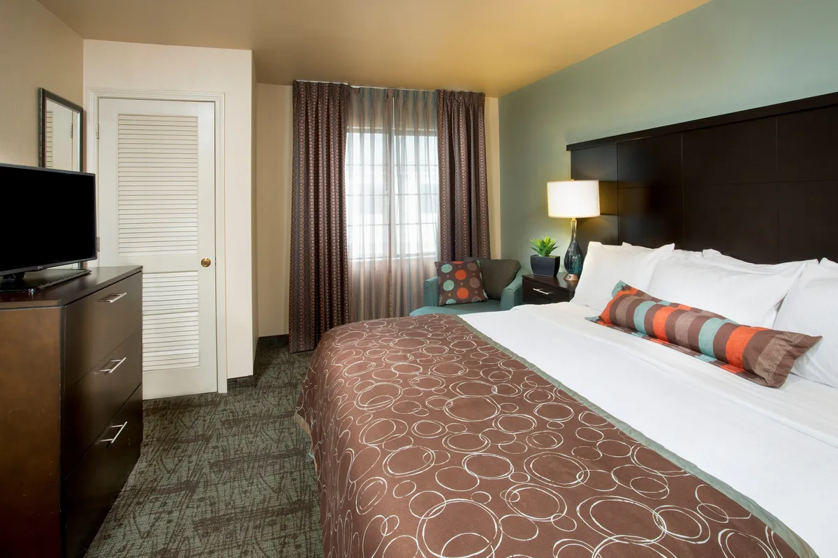 A welcoming suite interior featuring a comfortable bed with decorative pillows, a plush armchair, and a flatscreen TV situated across from the bed. Soft natural light filters through the curtains, creating a cozy and inviting atmosphere perfect for relaxation or enjoying a quiet evening in.