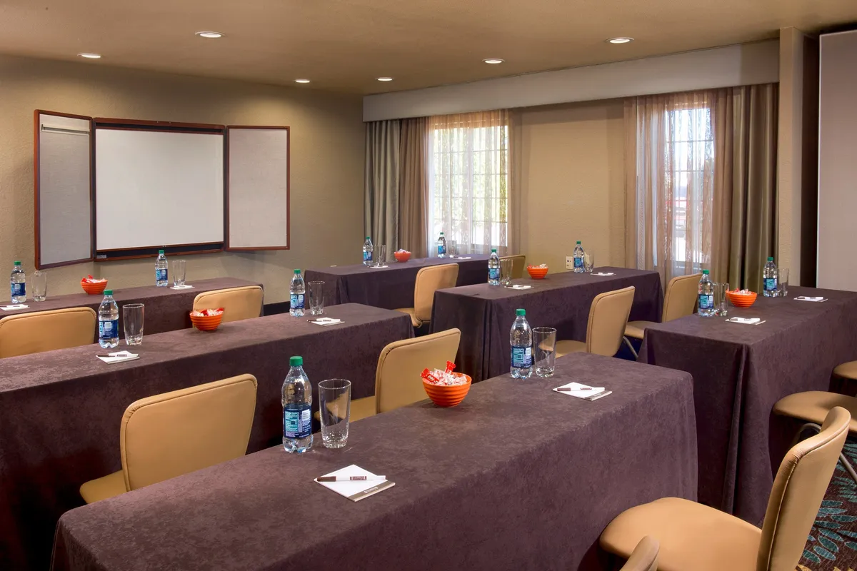 A spacious meeting room arranged with long tables, water bottles, snack bowls, and a presentation board, ideal for meetings, workshops, or collaborative sessions. The setup encourages productivity and group focus, with an atmosphere designed for professional gatherings.