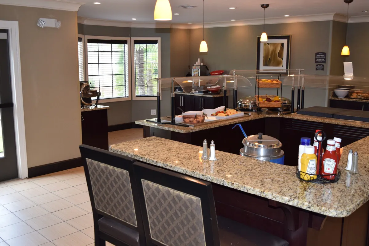 The image depicts an interior dining space with granite countertops, a well-displayed buffet, and ample seating. Guests are seen socializing and enjoying a hearty breakfast, with the space exuding a welcoming, communal vibe.