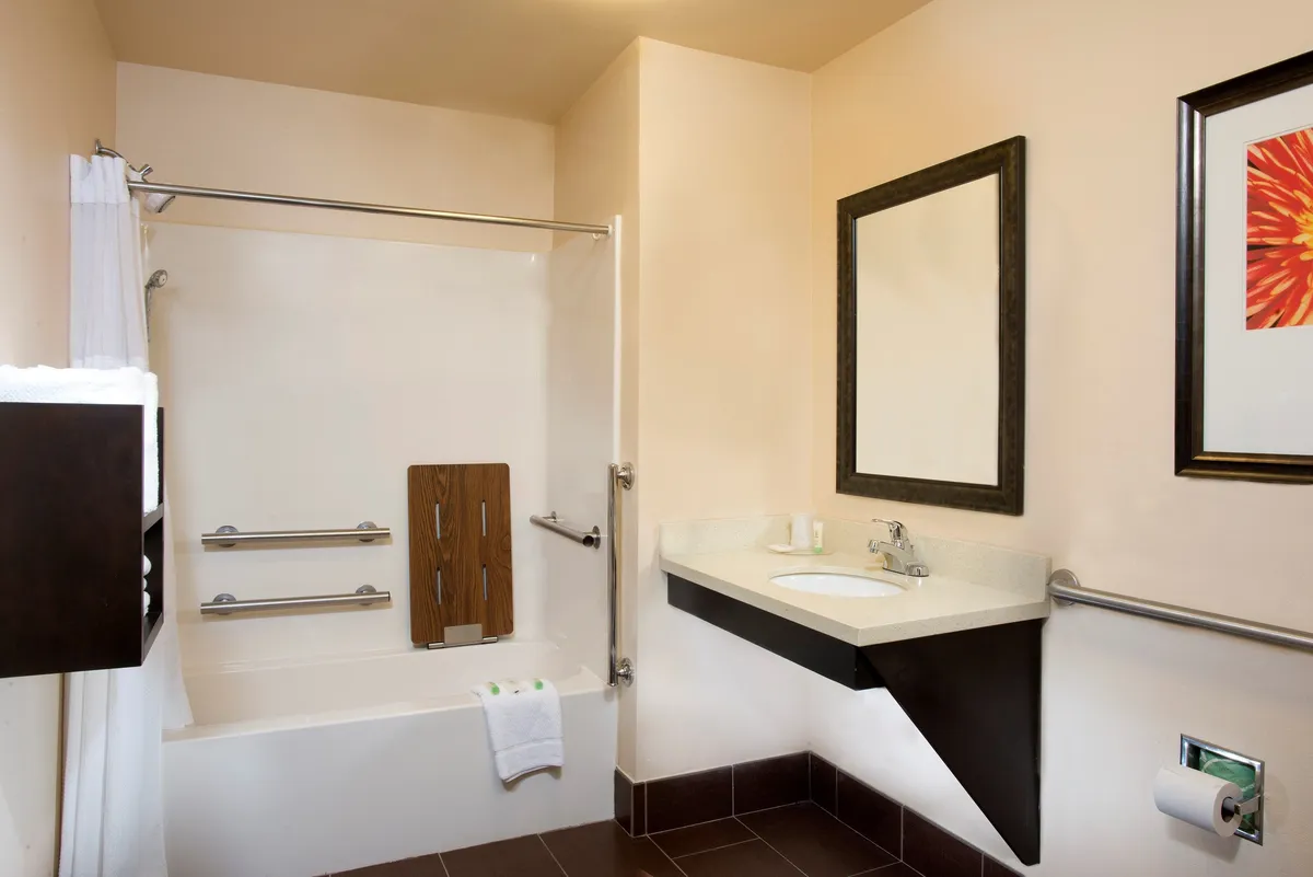 A serene bathroom interior featuring a bathtub with handrails, a modern sink, a large mirror, and tasteful wall art. The layout is open and accessible, with fixtures positioned for ease of use and comfort.
