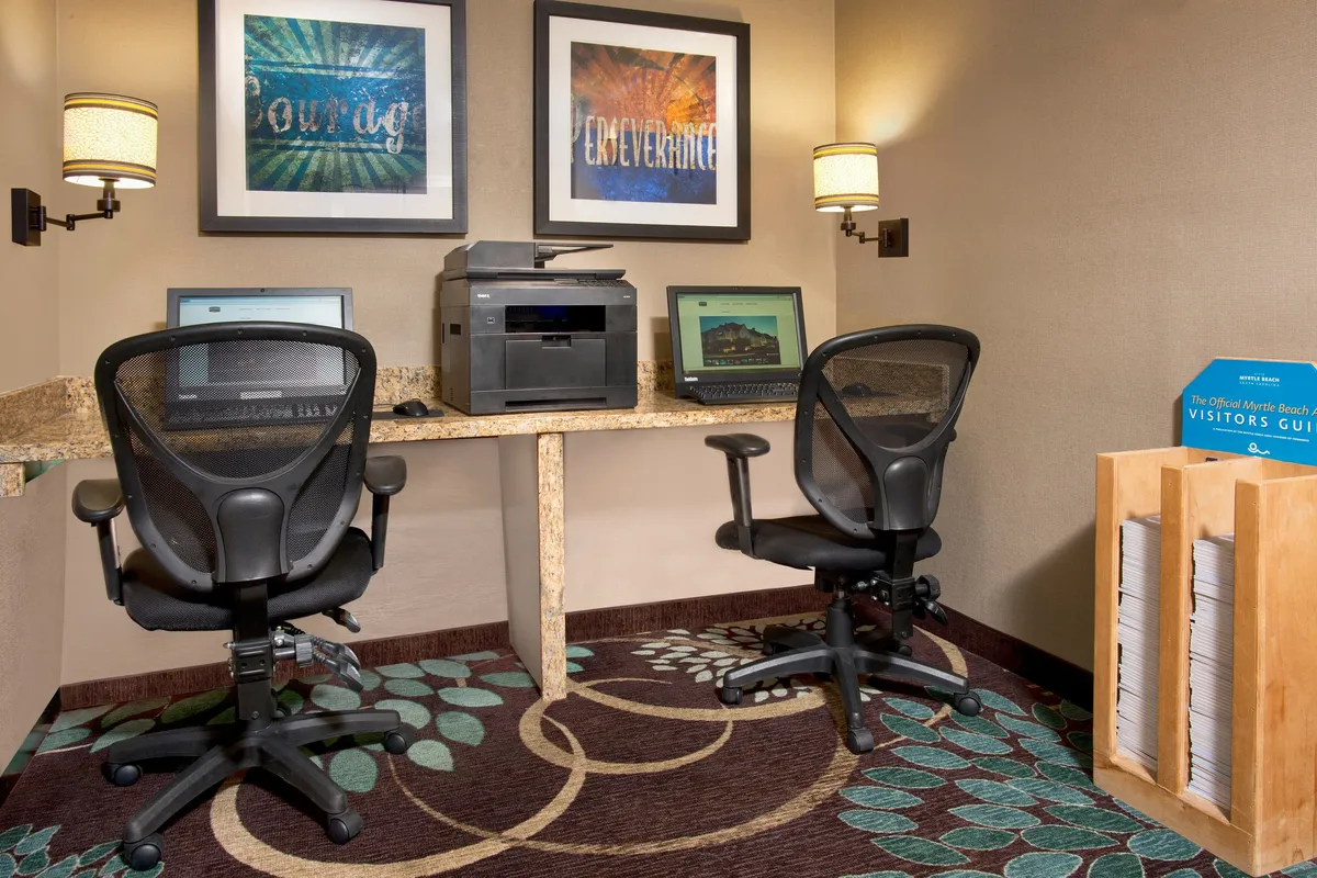 A modern business center with computer workstations, a printer, motivational artwork, and a magazine rack. The space is bright and organized, designed to support productivity and focused work for business travelers or guests who need to stay connected.