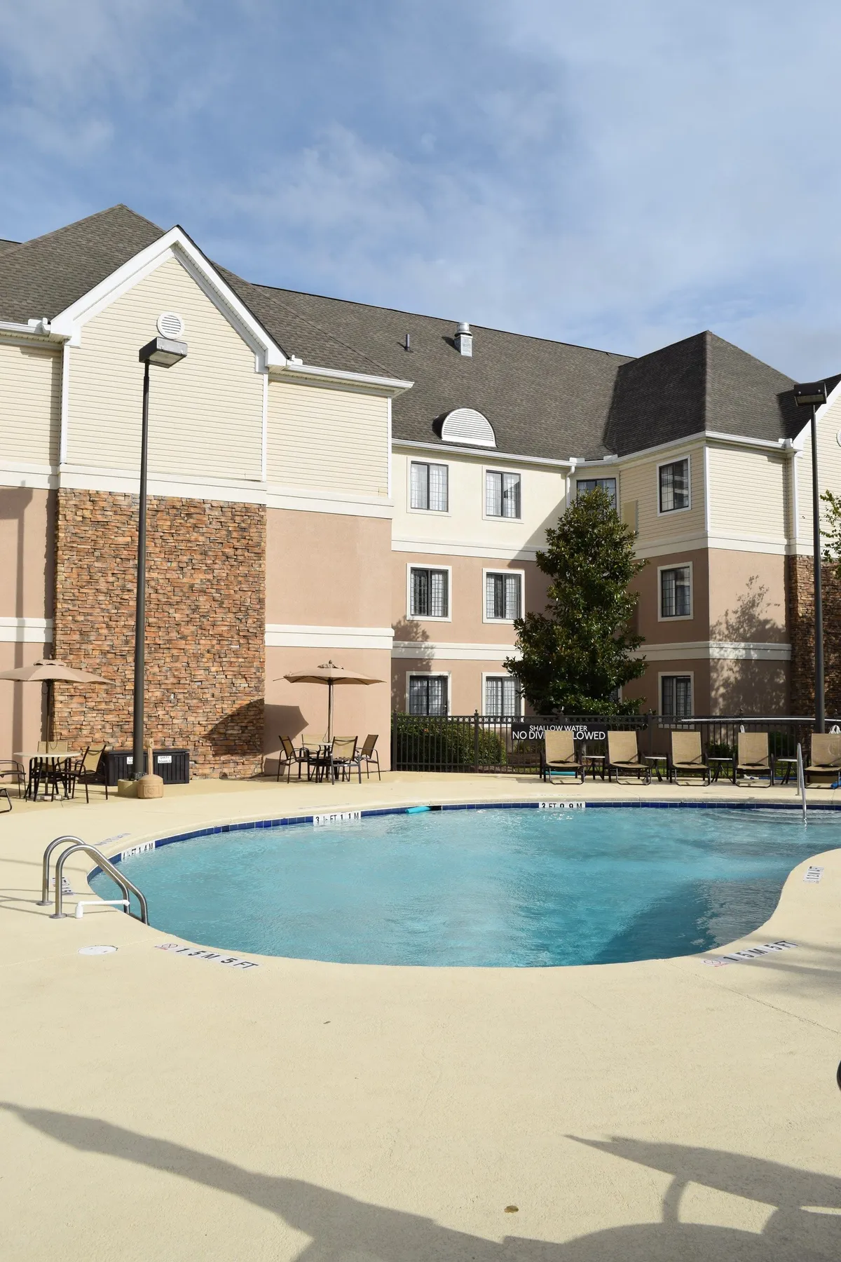 A bright, sunlit view of the swimming pool area with lounge chairs and umbrellas, adjacent to the building's brick wall. The scene captures families and couples enjoying a sunny day by the pool, making memories together in a recreational setting.