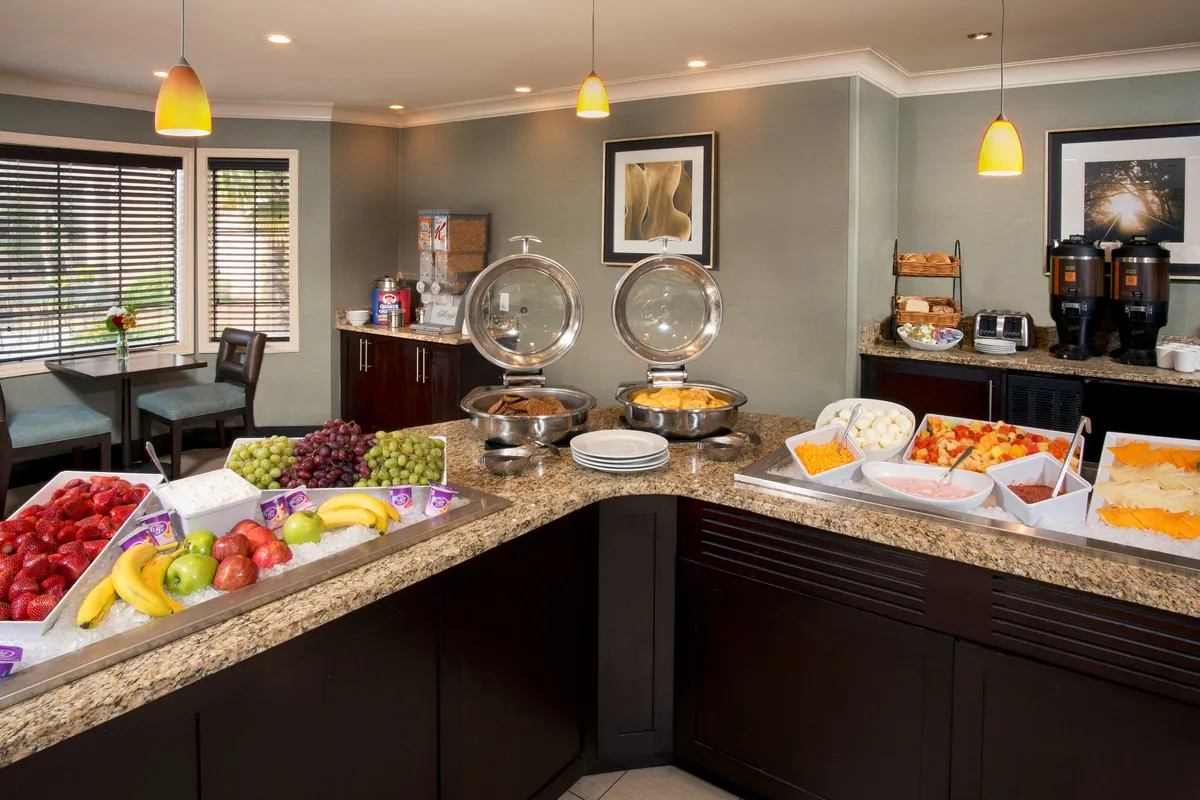 A cheerful breakfast area with a buffet table stocked with assorted fruits, chafing dishes displaying hot breakfast items, and a well-equipped coffee station. Guests are seen enjoying their morning meal in a bright, communal space.