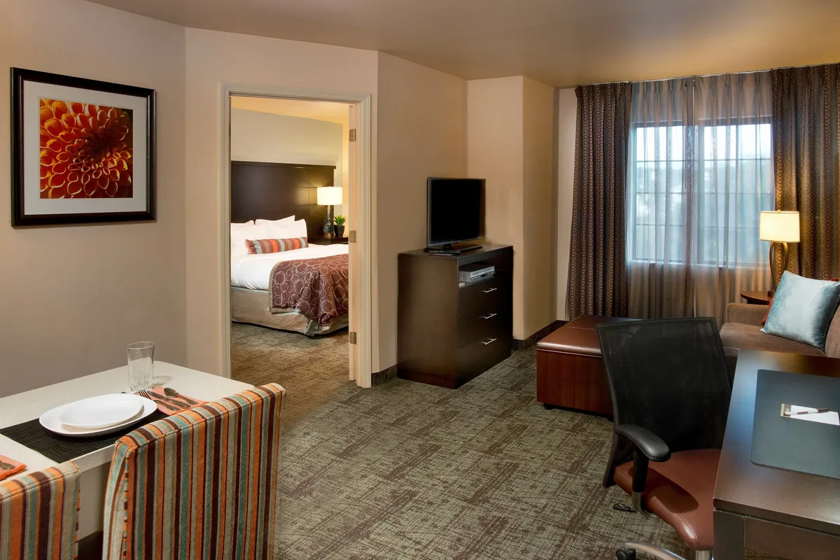 The image showcases the condo’s open and airy main living space featuring a modern dining table with chairs, a glimpse into the cozy bedroom through an open doorway, a sleek television unit for entertainment, and a sunlit window draped with soft curtains. The space is tastefully furnished, inviting guests to dine, relax on the sofa, catch up on work, or enjoy a movie night, all in a warm and comfortable atmosphere.
